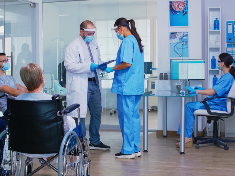 Doctor with face mask against covid19 discussing with nurse in hospital waiting area. Disabled senior woman in wheelchair waiting for examination. Assistant working on reception computer.